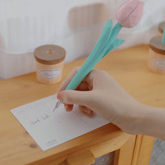 A person writing a "Good luck" message on a postcard using a pink flower pen.