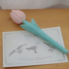 A pink flower pen lying on a wooden table beside a vintage-style postcard and a small pot.