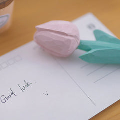 A pink flower pen resting on a wooden table next to a "Good luck" postcard.