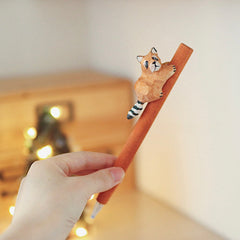 Hand holding a wooden pen with a hand-carved raccoon figure, with a softly lit background.