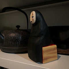 No Face Figure wooden sculpture sitting on a shelf beside a cast iron teapot.