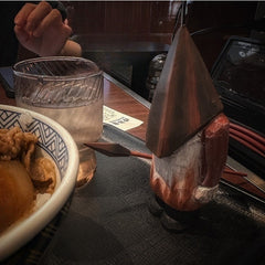 Pyramid Head wooden statue placed beside a meal and drink on a table, creating a surreal scene.