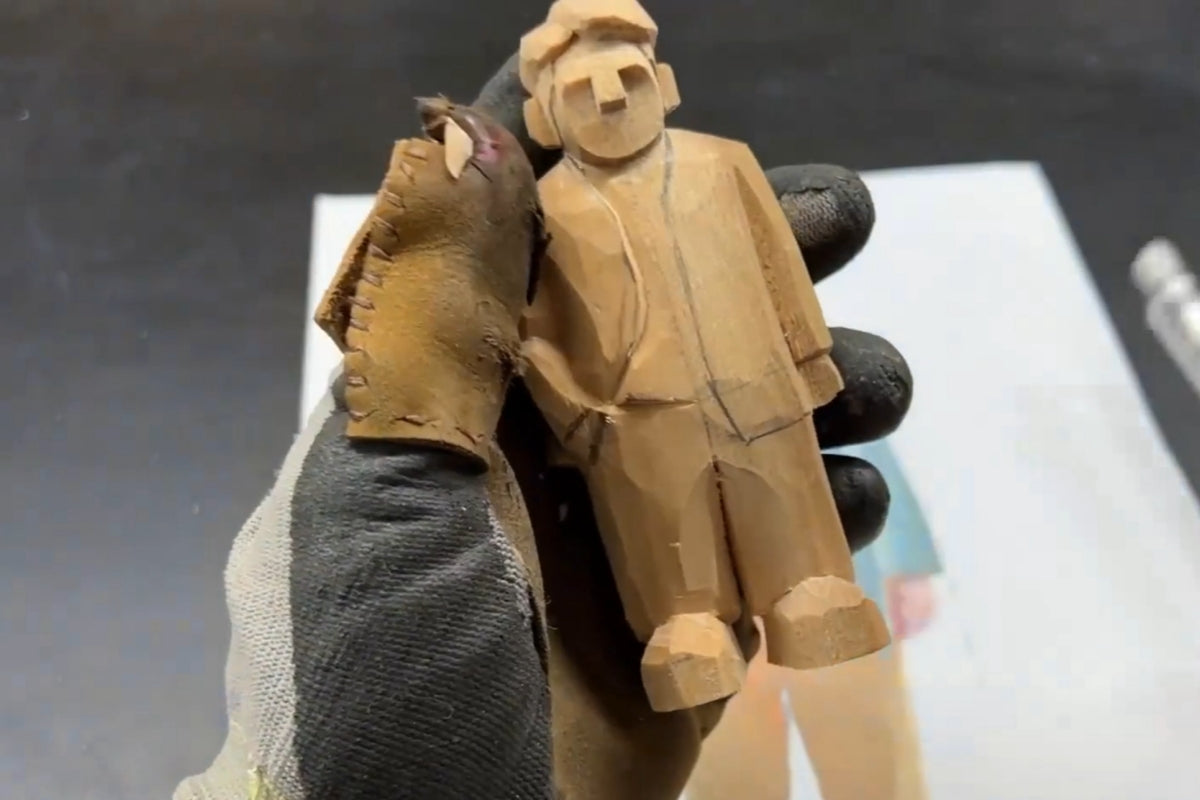 Front-facing wooden figure showing rough outlines of coat and pants with pencil sketch details for wood carving